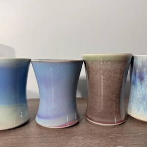 May include: Four handmade ceramic vases with a unique hourglass shape. Each vase features a different color gradient, including shades of blue, purple, and brown. The vases are displayed on a wooden surface.