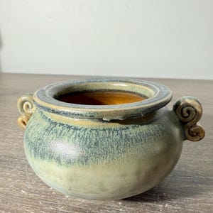 May include: A round ceramic bowl with a wide rim and two small, curled handles on opposite sides. The exterior features a mottled green and blue glaze, while the interior is finished in a smooth, warm brown tone with a glossy surface texture.