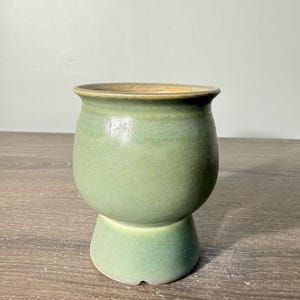 May include: A ceramic cup with a rounded body and a flared base, featuring a smooth matte finish in shades of sage green and olive. The interior rim shows a warm tan hue, and the vessel has a wide opening with a slightly curved lip.