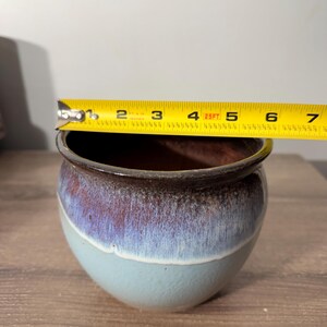 May include: A small, round ceramic pot with a diameter of approximately 5 inches. The pot features a gradient of colors, transitioning from a light blue base to a purple-brown rim. The interior is a dark brown color.