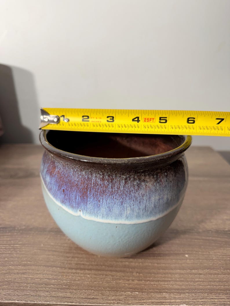 May include: A small, round ceramic pot with a diameter of approximately 5 inches. The pot features a gradient of colors, transitioning from a light blue base to a purple-brown rim. The interior is a dark brown color.