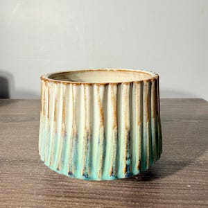 May include: A cylindrical ceramic planter featuring a vertical ribbed texture. The exterior displays a gradient color transition from a creamy off-white at the top rim to a vibrant turquoise blue at the base, with brown accents along the ridges.