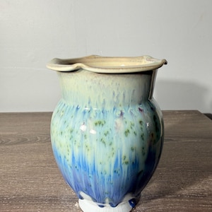 May include: A ceramic vase with a scalloped rim, featuring a glossy glaze that transitions from a creamy beige at the top to a vibrant blue at the base, with speckled green and purple accents throughout the body and a white footed base.