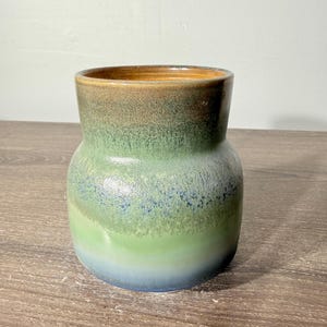May include: A ceramic vase featuring a gradient glaze that transitions from a deep brown rim to a vibrant green midsection, ending in a soft blue base. The vessel has a wide, rounded body that tapers slightly toward the top opening.