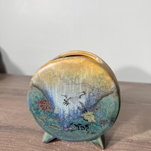 May include: A circular ceramic vase with a flat front featuring a painted scene of two cranes amidst colorful foliage and trees. The vase has a gradient finish transitioning from a golden yellow at the top to a deep teal and green at the base.