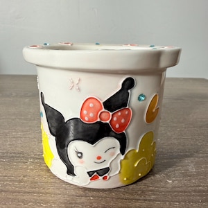 May include: A white ceramic planter featuring a raised, colorful graphic of a black and white character with a red polka-dot bow. The design includes yellow cloud-like shapes, a small crescent moon, and tiny blue rhinestones embedded on the rim.