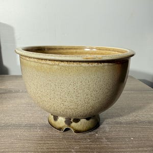 May include: A speckled, beige ceramic bowl with a wide rim and a pedestal base. The bowl has a rounded shape and a textured surface. The base features a brown, spotted pattern. The bowl is likely for decorative or functional use.