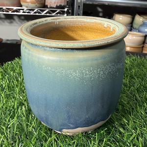 May include: A ceramic planter with a gradient blue glaze. The rim is a light green color, and the interior is a warm yellow. Other ceramic pots are visible in the background, suggesting a pottery theme.