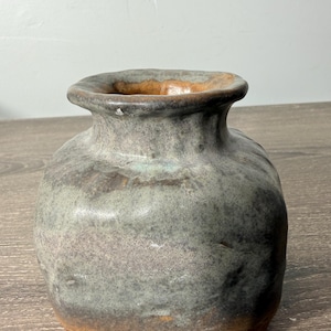 May include: A handcrafted ceramic vase with a rounded body and a flared rim. The vase features a mottled glaze in shades of gray, green, and brown. The base has three small feet. This decorative piece is suitable for displaying flowers or as a standalone art object.