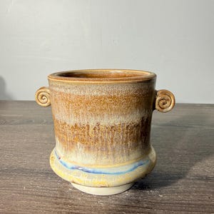 May include: A ceramic vessel featuring a cylindrical body with two small, curled handles on the sides. The exterior displays a mottled glaze in shades of tan, brown, and cream, with a distinct blue horizontal band near the flared base.