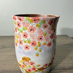May include: A tall, ceramic vase with a floral design. The vase features pink flowers, green leaves, and a brown branch against a white and lavender background. The rim is brown.