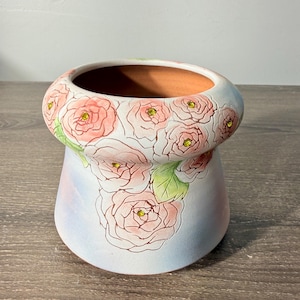 May include: A ceramic planter featuring a hand-painted design of soft pink roses with green leaves against a light blue background. The center of each rose is accented with a small, sparkling yellow rhinestone embedded into the ceramic surface.