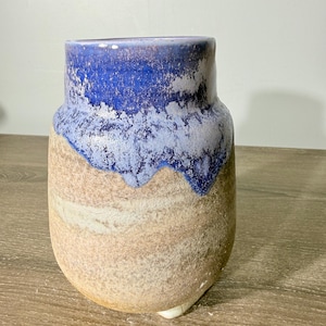 May include: A ceramic vase with a rounded body and a wide opening. The upper portion features a vibrant blue glaze that drips down over a textured, matte, sandy-beige base. The surface shows a distinct contrast between the smooth, glossy blue top and the earthy, granular lower section of the vessel.