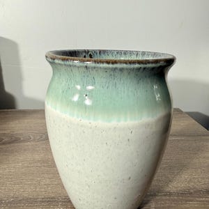 May include: A handcrafted ceramic vase with a two-tone design. The top half is a seafoam green, transitioning to a speckled off-white base. The rim has a dark brown accent. This decorative vase is suitable for home decor.
