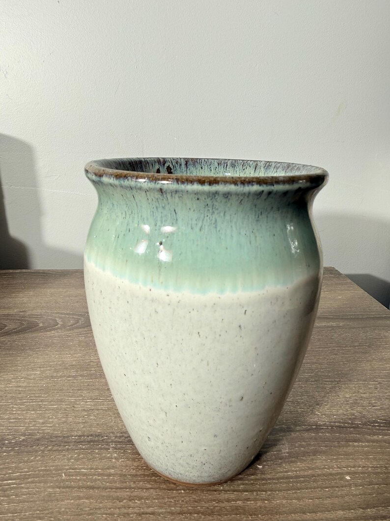 May include: A handcrafted ceramic vase with a two-tone design. The top half is a seafoam green, transitioning to a speckled off-white base. The rim has a dark brown accent. This decorative vase is suitable for home decor.