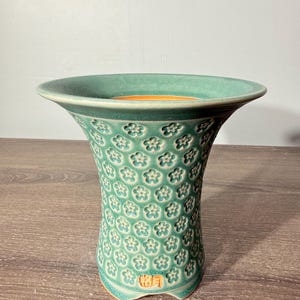 May include: A flared ceramic vase with a matte teal green exterior featuring a repeating embossed flower pattern. The interior of the vase is a contrasting warm orange hue. Near the base, there is a rectangular stamp with two Japanese characters.