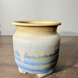 May include: A ceramic pot with a wide rim and three small feet. The upper portion features a smooth tan glaze, while the lower section displays a textured blue and tan drip pattern that cascades down the sides of the rounded vessel body.