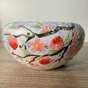 May include: A rounded ceramic bowl featuring a hand-painted design of flowering branches with pink blossoms, green leaves, and red fruit. The surface is decorated with small white paint splatters and embedded green and pink rhinestones.