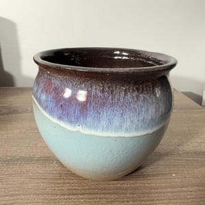 May include: A handcrafted ceramic bowl with a rounded shape. The bowl features a gradient of colors, transitioning from a deep brown rim to a purple-blue midsection, and ending with a light blue base. The bowl is likely used for decorative or functional purposes.