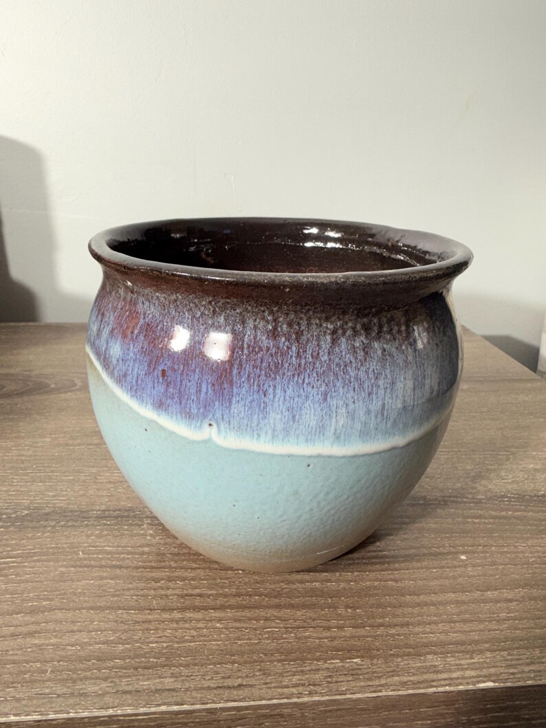 May include: A handcrafted ceramic bowl with a rounded shape. The bowl features a gradient of colors, transitioning from a deep brown rim to a purple-blue midsection, and ending with a light blue base. The bowl is likely used for decorative or functional purposes.