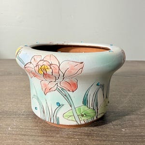 May include: A ceramic planter with a wide, flared rim and a light blue glaze. The exterior features hand-painted pink lotus flowers, green lily pads, and thin green stems. Small, round, blue rhinestones are embedded along the rim and body.