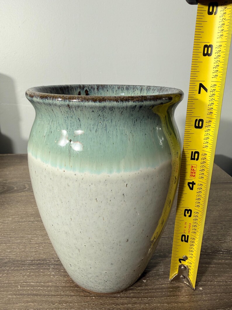 May include: A handcrafted ceramic vase with a two-tone glaze. The top portion is a seafoam green, transitioning to a speckled off-white base. The vase is approximately 7 inches tall and has a wide opening.