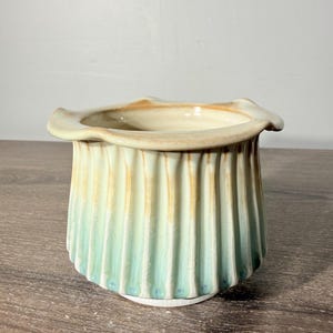May include: A ceramic planter with a fluted, vertical ribbed exterior featuring a gradient glaze that transitions from a soft mint green at the base to a warm, toasted tan at the scalloped, flared rim, showcasing a smooth, glossy finish.