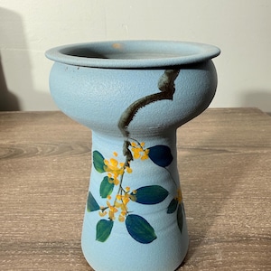 May include: A light blue ceramic vase with a bulbous top and a slender base. The vase is decorated with a floral design featuring yellow flowers and green leaves. A dark brown line runs down the side of the vase. The vase is sitting on a wooden surface.
