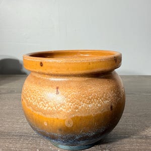 May include: A round ceramic pot featuring a gradient glaze that transitions from a warm golden-orange at the wide rim to a speckled tan in the center, ending in a deep blue-grey base. The vessel has a thick, rounded lip and a smooth finish.