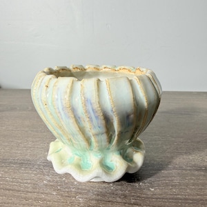 May include: A small ceramic bowl with a fluted, scalloped rim and a ruffled base. The exterior features a pale mint green glaze with a subtle crackle finish and brown accents along the vertical ridges, creating a textured, shell-like appearance.