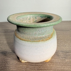 May include: A ceramic pot with a wide, flat rim featuring a green glaze that transitions into a light, off-white base. The vessel has a rounded body and sits on three small, short feet, showcasing a textured, speckled finish throughout the surface.