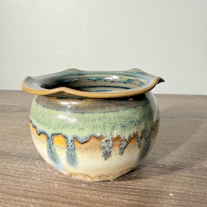 May include: A round ceramic bowl with a wide, wavy rim. The exterior features a reactive glaze with layers of sage green, deep blue, and warm tan, creating a dripping effect that transitions into a creamy off-white base at the bottom.
