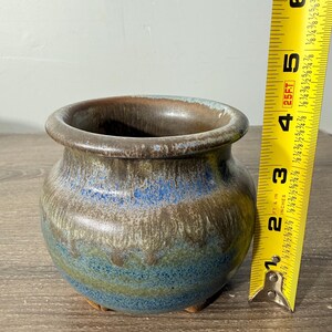 May include: A small, handcrafted ceramic pot with a wide opening. The pot features a blend of blue, brown, and green hues. The pot is approximately 3 inches tall and 4 inches wide.