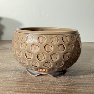 May include: A round ceramic bowl with a muted blue-grey glaze and a repeating pattern of impressed sunburst or flower motifs. The bowl features a wide opening, a curved body, and a footed base with a small rectangular cutout near the bottom edge.