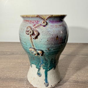 May include: A ceramic vase with a flared rim and a bulbous body featuring a crackle glaze finish. The surface displays a gradient of colors including purple, teal, and white, with decorative raised floral-like elements and blue drip accents.