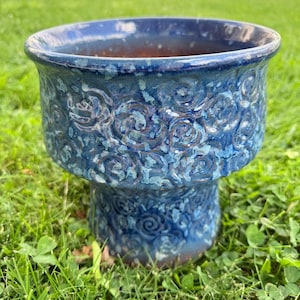 May include: A blue ceramic planter with a pedestal base. The planter has a wide, rounded rim and is decorated with a repeating floral pattern. The planter is sitting on a bed of green grass.