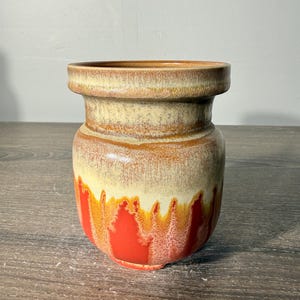 May include: A ceramic vase with a wide, flat rim and a rounded body. The upper portion features a smooth, creamy beige glaze, while the lower section displays a vibrant, dripping red and orange glaze that creates a textured, layered appearance.