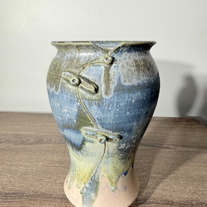 May include: A ceramic vase with a flared rim and a tapered base, featuring a glaze in shades of blue, green, and cream. The exterior surface is decorated with a raised, linear design resembling toggle closures that wrap around the upper body.