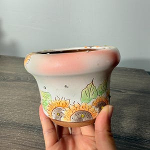 May include: A ceramic flower pot with a flared rim, featuring a hand-painted sunflower design with green leaves on a white base. The upper rim is shaded with a soft pink hue, and the exterior is embellished with small, clear, reflective rhinestones.