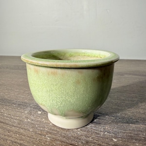 May include: A ceramic bowl with a wide rim and a pedestal base. The exterior features a mottled light green glaze with subtle brown accents, while the interior and base are a smooth, off-white color with a glossy finish.