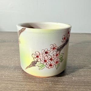 May include: A cylindrical ceramic planter featuring a hand-painted design of a brown tree branch with light pink cherry blossoms and green leaves. The center of each flower is accented with a small, sparkling red rhinestone embedded in the clay.