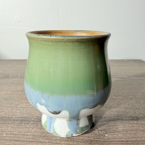 May include: A ceramic cup featuring a smooth, glossy finish with a color gradient transitioning from a deep green at the rim to a light blue in the middle, ending in a white, crackled glaze texture at the base with a flared pedestal foot.