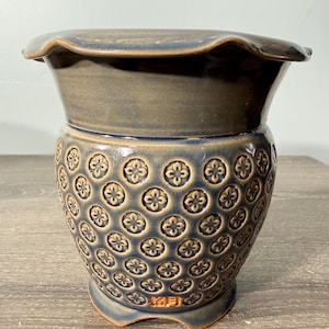 May include: A blue and tan ceramic planter with a ruffled rim. The body of the planter is decorated with a repeating floral pattern. The planter has a wide opening and a sturdy base.