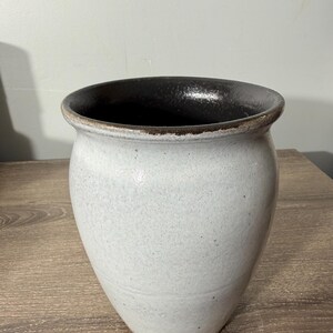 May include: A handcrafted, off-white ceramic vase with a dark brown interior and rim. The vase has a wide opening and a rounded, tapered body. The exterior has a speckled texture, and the vase sits on a wooden surface.