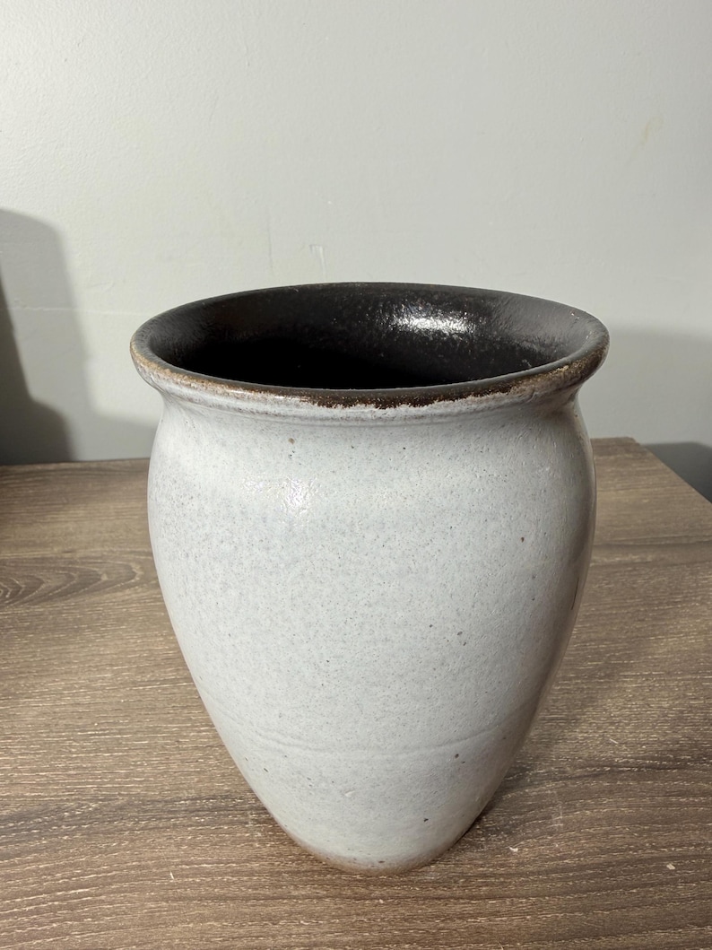 May include: A handcrafted, off-white ceramic vase with a dark brown interior and rim. The vase has a wide opening and a rounded, tapered body. The exterior has a speckled texture, and the vase sits on a wooden surface.