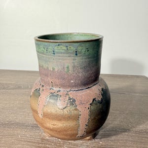 May include: A ceramic vase with a rounded base and a wide neck, featuring a multi-colored glaze. The upper section is a deep green, while the lower body transitions into shades of brown and tan with a textured, dripping pinkish-mauve glaze.