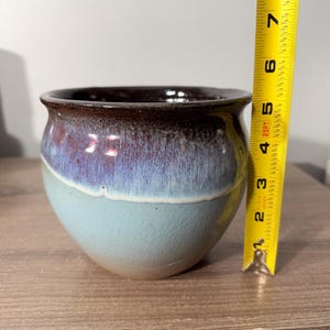 May include: A handcrafted ceramic pot with a rounded shape and a wide opening. The pot features a gradient of colors, transitioning from a dark brown rim to a purple-blue midsection and a light blue base. The pot measures approximately 4 inches in height.