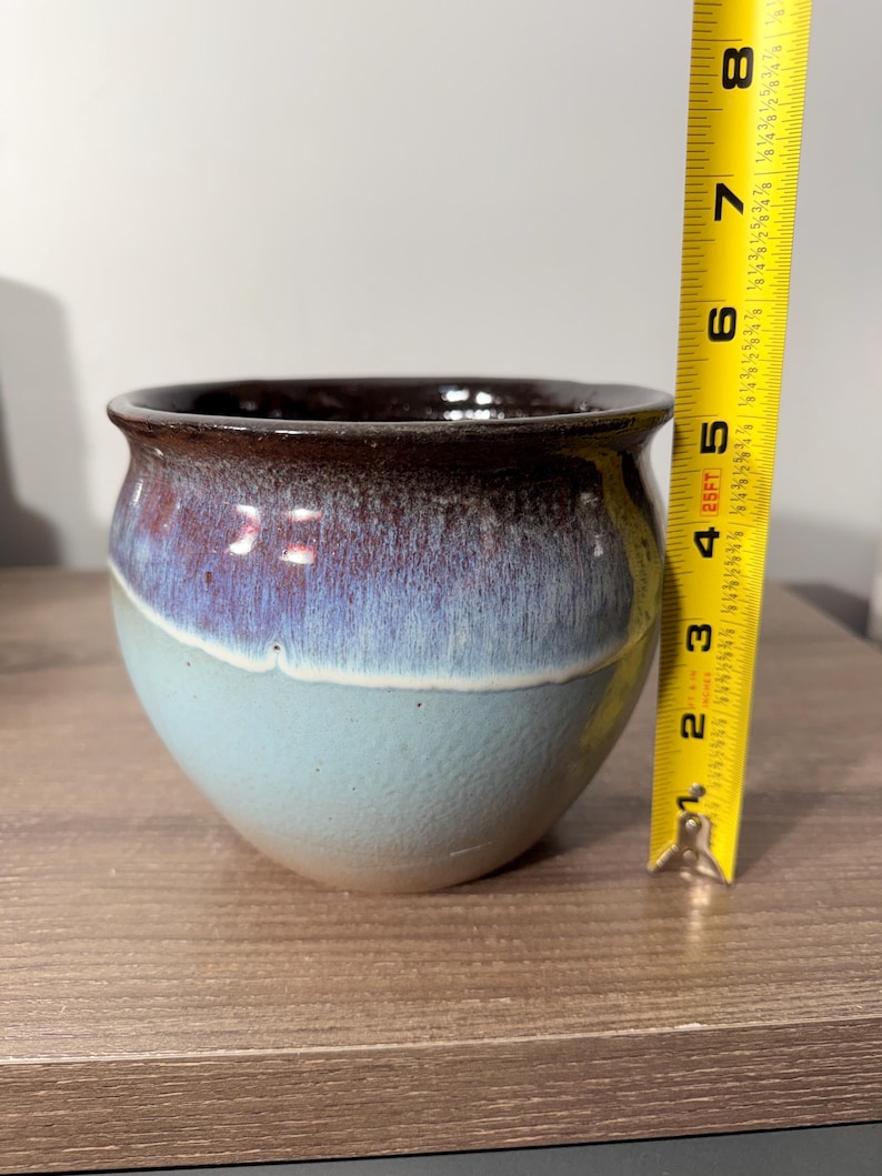 May include: A handcrafted ceramic pot with a rounded shape and a wide opening. The pot features a gradient of colors, transitioning from a dark brown rim to a purple-blue midsection and a light blue base. The pot measures approximately 4 inches in height.