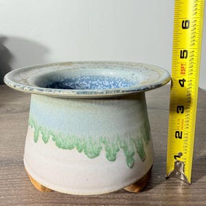 May include: A ceramic planter with a wide rim, featuring a white base, a green drip glaze pattern, and a blue interior. A yellow measuring tape placed vertically next to the planter indicates a height of approximately 4.25 inches.