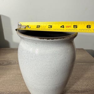 May include: A handmade, off-white ceramic vase with a dark brown rim. The vase is approximately 6 inches tall and has a speckled texture. The vase is sitting on a wooden surface.